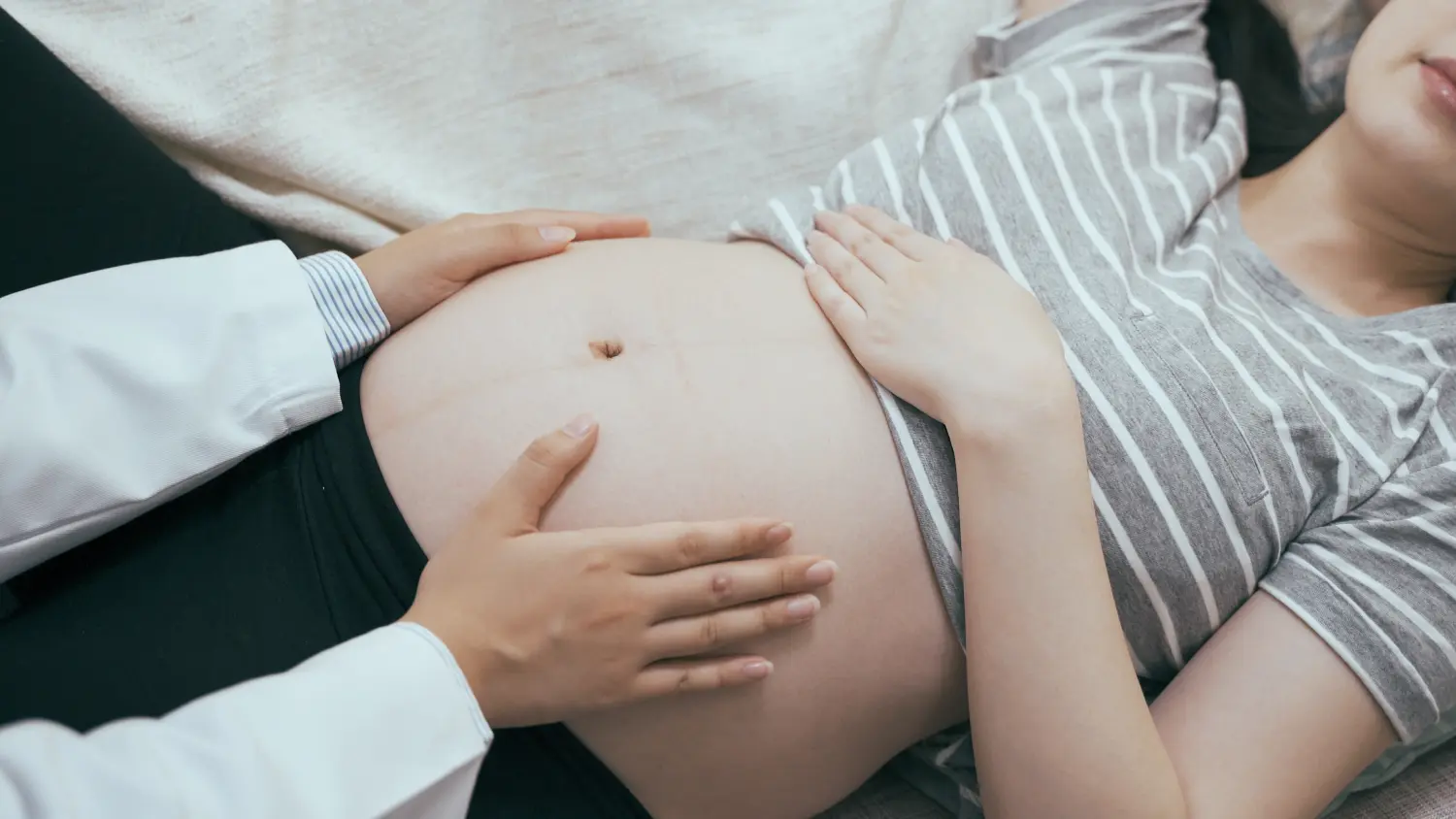 Hebamme überprüft den Herzschlag und die Bewegung des Babys, während sie der schwangeren Frau die Hände auf den nackten Bauch legt. Unbekanntes weibliches medizinisches Personal in weißem Gewand, das sich berührt