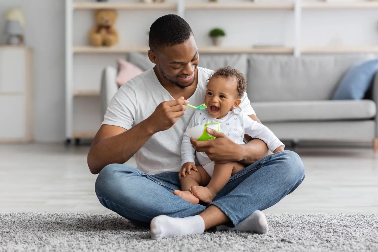 Glücklicher junger schwarzer Papa füttert seinen entzückenden kleinen Sohn zu Hause mit einem Löffel