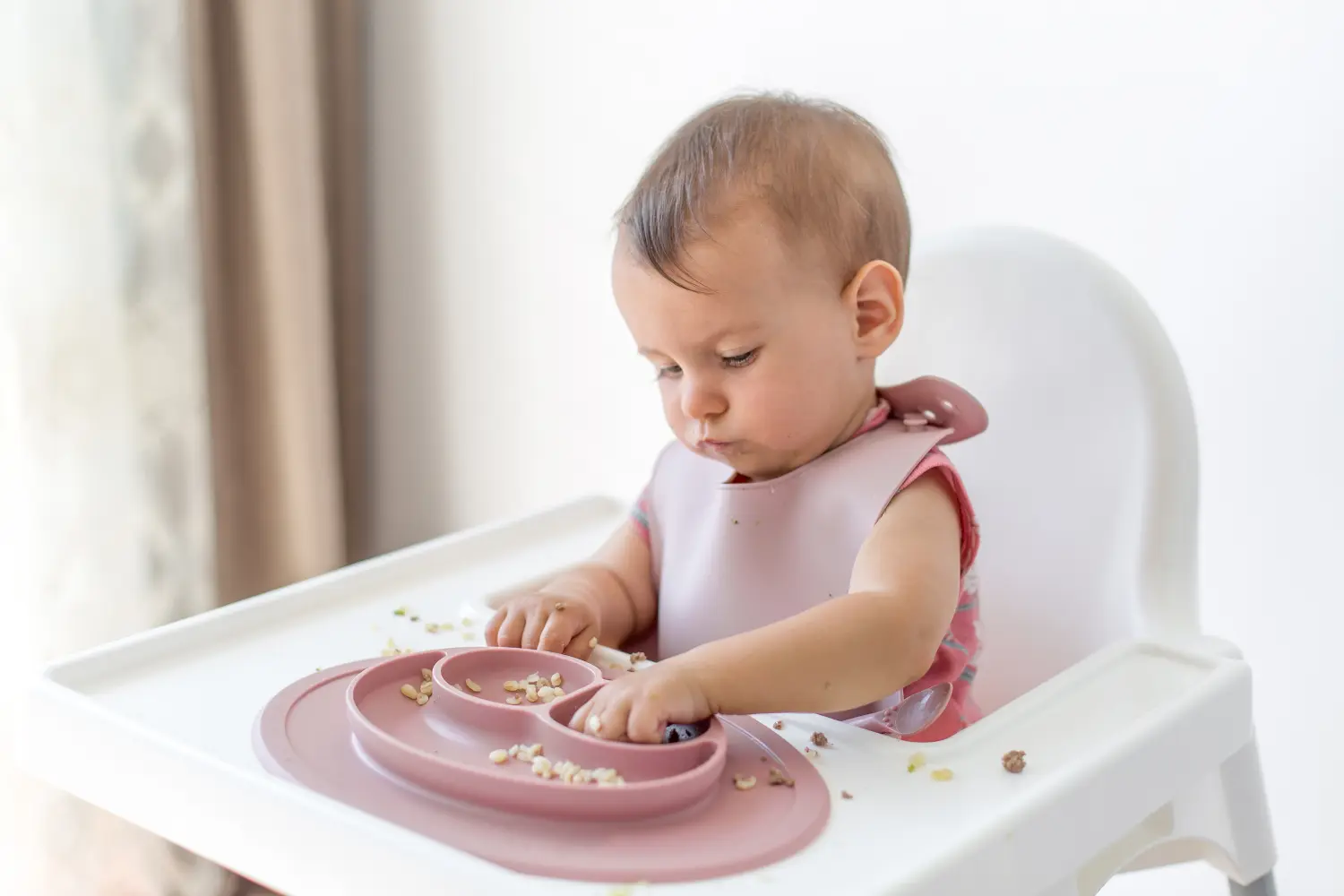 Ein 8 Monate altes kleines Mädchen sitzt in einem Hochstuhl und isst Beikost, Bulgur, Müsli und Fleisch, Nahaufnahmeporträt blickt in die Kamera.